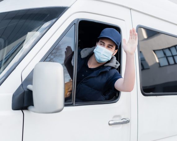 Courier in medical mask waving hand while driving auto