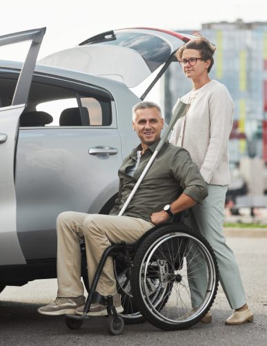 Full length portrait of mature couple with man in wheelchair by car in parking lot outdoors, copy space